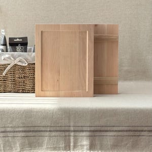 May include: Two unfinished wooden cabinet doors, one with a recessed panel, stand upright on a neutral-toned surface. The wood has a light, natural colour. A woven basket with a white bow is partially visible in the background.