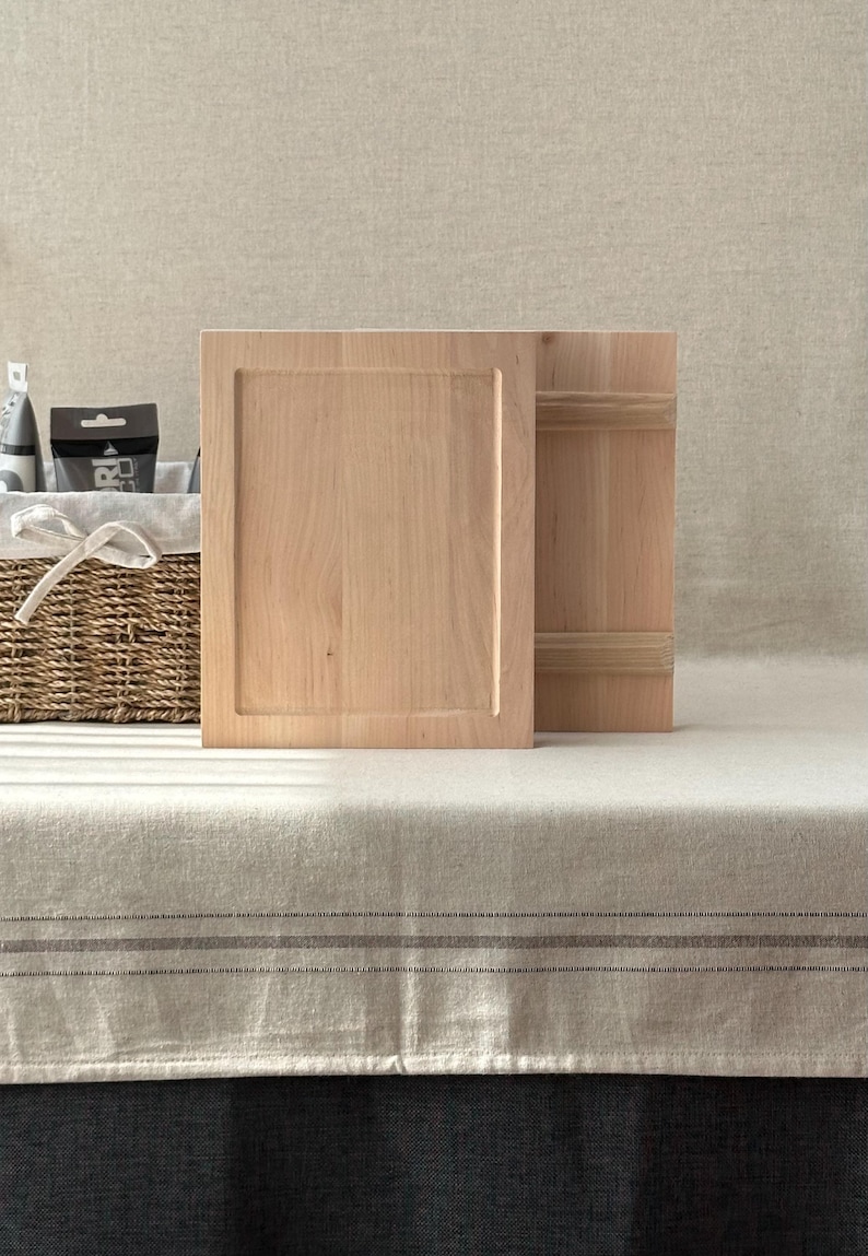 May include: Two unfinished wooden cabinet doors, one with a recessed panel, stand upright on a neutral-toned surface. The wood has a light, natural colour. A woven basket with a white bow is partially visible in the background.