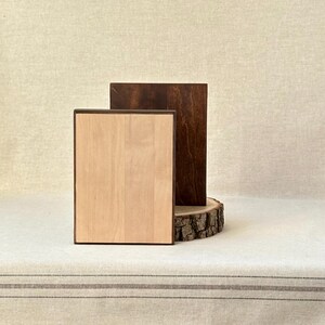 May include: Two wooden rectangular plaques. One is light-coloured with a dark brown frame, and the other is dark brown. They are displayed on a light-coloured surface, with the dark brown plaque resting on a wooden slice.
