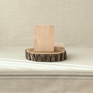 May include: Two light-colored wooden blocks, one standing upright and the other lying down, are displayed on a rustic, circular wooden slice. The wood slice has a natural bark edge. The background is a neutral-toned fabric.