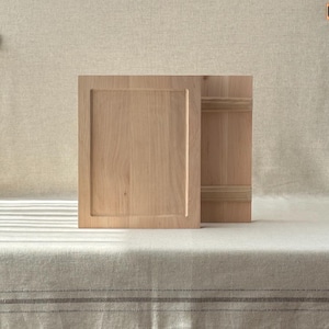 May include: Two light-coloured wooden cabinet doors, one open and one closed, with a recessed panel design. The doors are set against a neutral background, suggesting a minimalist or rustic aesthetic. The wood grain is visible.
