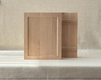 May include: Two light-colored wooden cabinet doors, one open and one closed, with a recessed panel design. The doors are set against a neutral background, suggesting a minimalist or rustic aesthetic. The wood grain is visible.