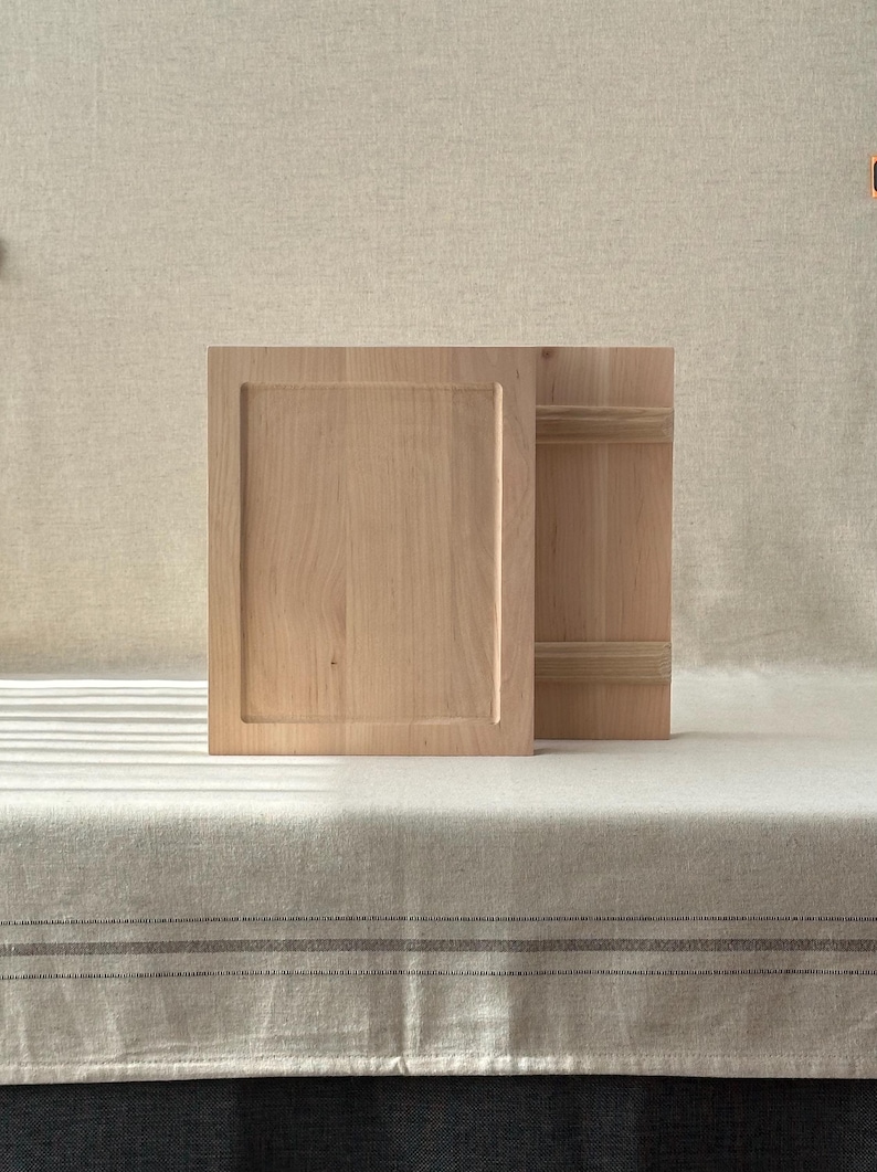 May include: Two light-coloured wooden cabinet doors, one open and one closed, with a recessed panel design. The doors are set against a neutral background, suggesting a minimalist or rustic aesthetic. The wood grain is visible.
