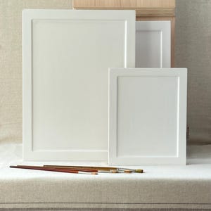 May include: Three white framed canvases of varying sizes are arranged on a neutral-toned surface. Several paintbrushes with wooden handles are placed in front of the canvases. A wooden box is visible in the background.