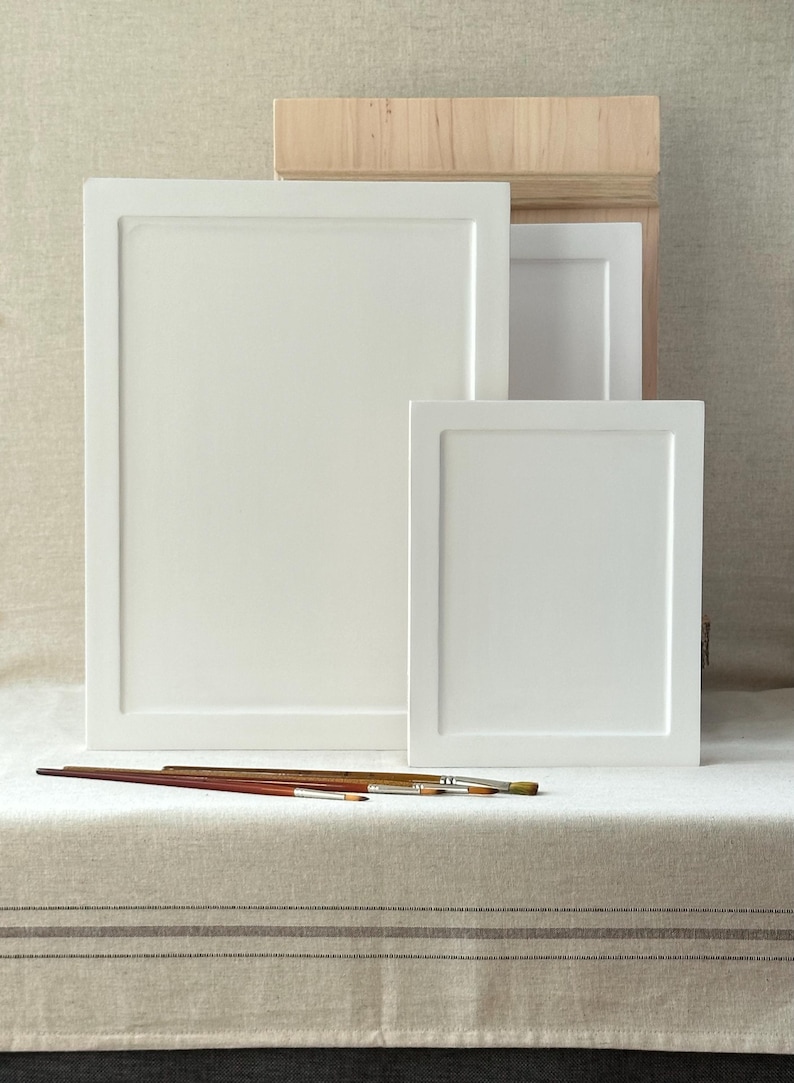May include: Three white framed canvases of varying sizes are arranged on a neutral-toned surface. Several paintbrushes with wooden handles are placed in front of the canvases. A wooden box is visible in the background.