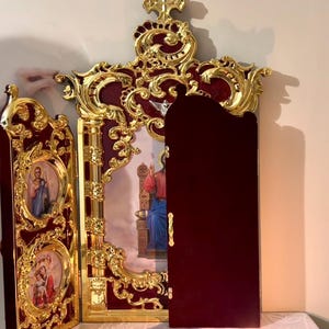 Pode incluir: Ícone religioso ornamentado, com moldura dourada e fundo bordô. O ícone apresenta figuras religiosas e é decorado com intrincados filigranas dourados. Uma vela acesa e um prato pequeno com comida estão em primeiro plano.