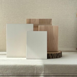 May include: A studio shot featuring a collection of blank canvases and wooden blocks. Two white rectangular canvases stand in front of two wooden blocks and a wood slice. The canvases are different sizes. The background is a neutral beige.