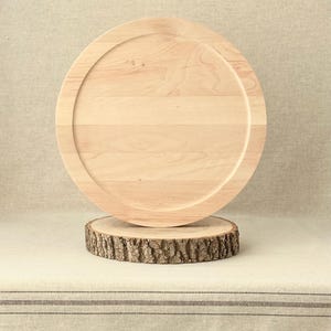 May include: A light-coloured wooden serving tray with a raised edge, resting on a small, round wooden base made from a tree slice. The tray and base are displayed on a neutral-toned surface.
