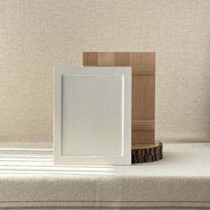May include: A white, square cabinet door with a recessed panel stands next to a wooden panel and a wood slice. The cabinet door is painted a solid, matte white. The wooden panel has a natural wood grain.