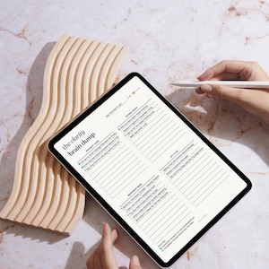 May include: A digital tablet displaying a planner page titled "the clarity brain dump" with various sections for tasks and notes. A wooden, wavy decorative piece is on the left. A stylus hovers over the screen.