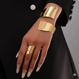May include: Gold-toned jewelry set featuring a wide cuff bracelet, a smaller bracelet, and a statement ring. The pieces have a smooth, polished finish and a modern, geometric design. The jewelry is displayed against a black background.