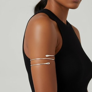 May include: Silver arm cuff featuring a three-band design. The bands wrap around the upper arm, with teardrop-shaped ends. The jewelry is worn on a person wearing a black sleeveless top.