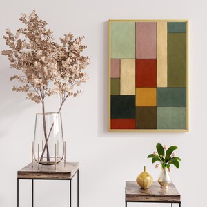 May include: Abstract geometric art print with a gold frame. The artwork features a collage of colorful rectangular shapes in shades of green, pink, red, yellow, and black. The print is displayed on a white wall, with decorative vases and a plant in the foreground.