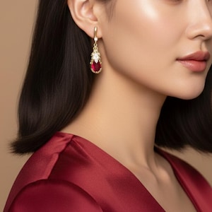 May include: Gold-toned drop earrings featuring a pear-shaped, deep red gemstone. The earrings have a floral design at the top with small, clear stones. The setting is gold, and the earrings are worn on a person's ear.