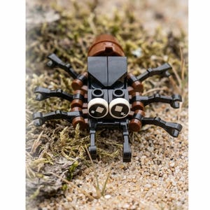 Puede incluir: Una pequeña figura con forma de insecto construida con bloques de construcción de plástico negro y marrón. La figura presenta una parte trasera redondeada de color marrón oscuro, un cuerpo central negro y ocho patas negras articuladas. Dos grandes ojos circulares blancos con pupilas cuadradas negras están situados en la parte delantera.