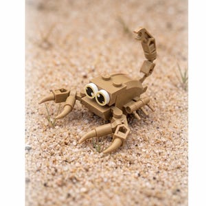 May include: A tan-colored scorpion figure constructed from plastic building blocks. The scorpion features two large front pincers, a curved tail with a claw-like tip, and two prominent, round white eyes with black pupils positioned on the front.