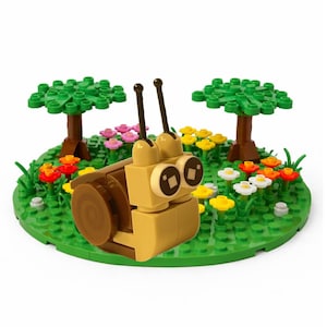 May include: A tan and brown snail made of toy blocks, set in a green grassy scene with colorful flowers and two green trees. The snail has large, round eyes and long antennae. The scene is on a green circular base.