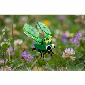 Pode incluir: Uma pequena figura de inseto construída com blocos de montar de plástico nas cores verde, amarelo e marrom. A figura apresenta grandes olhos brancos com pupilas pretas, asas verdes translúcidas com padrão de grade e pequenas pernas marrons.