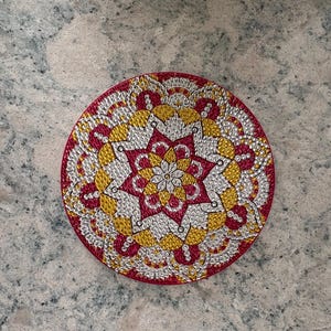May include: A round coaster with a mandala design. The coaster features a red border and a central starburst pattern in red, yellow, and silver. The design is made of small, sparkling elements.