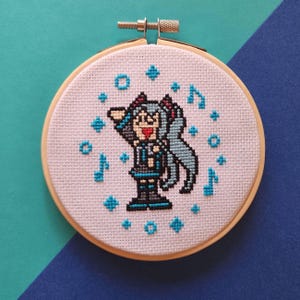 May include: A cross-stitch embroidery hoop featuring a cartoon character with turquoise hair and accents. The character is surrounded by musical notes and star-shaped designs in turquoise. The embroidery is set in a wooden hoop.