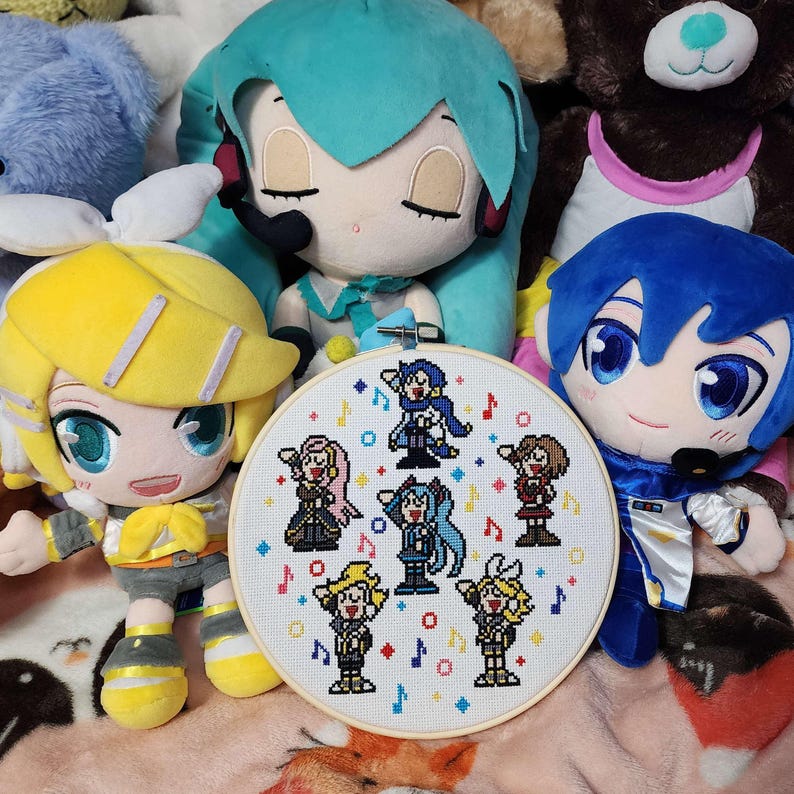 May include: A cross-stitch embroidery hoop with a pixelated design of anime characters and musical notes. The hoop is surrounded by plush dolls with blue, yellow, and teal hair. The dolls have large eyes and are wearing outfits.
