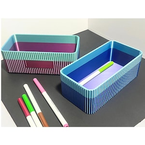May include: Two rectangular storage boxes with vertical striped sides and colour gradients. One box has purple, teal, and magenta hues, while the other features blue and purple tones. Several colourful markers are placed in front of the boxes.