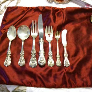 Könnte beinhalten: Eine Sammlung antiker Silberwaren, die auf einem tiefroten Samttuch angeordnet sind. Das Set umfasst Löffel, Gabeln, ein Messer und ein Buttermesser. Das Besteck hat verzierte florale Details an den Griffen.