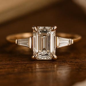 May include: A gold engagement ring with a large, rectangular emerald-cut diamond as the centerpiece. Two smaller trapezoid-cut diamonds flank the main stone. The ring is set on a gold band, displayed on a wooden surface.