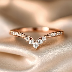 Può includere: Anello in oro rosa con un design curvo, incastonato con più piccole pietre preziose trasparenti. L'anello presenta un gruppo centrale di pietre più grandi, creando un aspetto delicato ed elegante. L'anello è esposto su un tessuto morbido e chiaro.