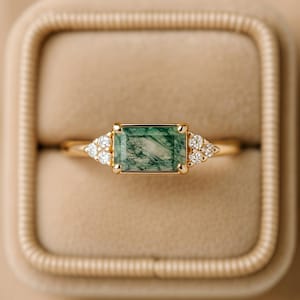Puede incluir: Un anillo de oro con una piedra rectangular de ágata musgosa verde. El anillo presenta pequeños diamantes redondos a cada lado de la piedra principal, engastados en una banda de oro. El anillo se muestra en un joyero beige.