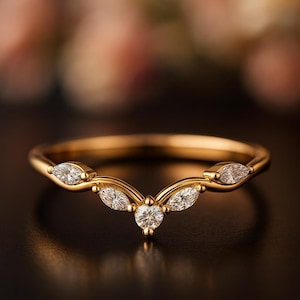 Może przedstawiać: Złoty pierścionek o zakrzywionym kształcie, z pięcioma przezroczystymi, błyszczącymi kamieniami szlachetnymi. Centralny kamień jest okrągły, a pozostałe mają kształt markizy. Pierścionek jest umieszczony na ciemnym tle.