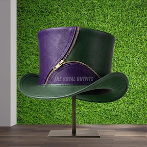 Puede incluir: Un sombrero de copa bicolor de cuero verde y morado, con un detalle de cremallera dorada. El sombrero se exhibe sobre un soporte metálico sobre un fondo de hierba verde. El texto "THE ROYAL OUTFITS" es visible en el sombrero.
