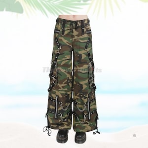 Può includere: Pantaloni cargo con stampa mimetica con più tasche, cerniere e catene. I pantaloni hanno cinghie nere e hardware argentato. Il design a gamba larga presenta una vita con coulisse e sono indossati con stivali neri.
