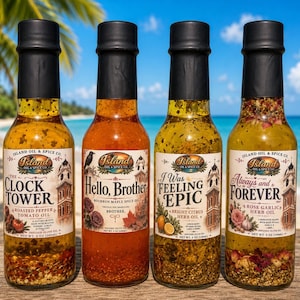 May include: Four glass bottles of Island Oil & Spice Co. infused olive oil. The labels read "The Clock Tower", "Hello, Brother", "I Was Feeling Epic", and "Always and Forever". Each bottle has a black cap and contains visible herbs and spices.