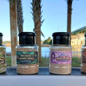 May include: Four clear spice jars with black lids are displayed outdoors. The jars are labeled "Dill Pickle Addiction," "Wicked Caramel Apple," "KPop Korean BBQ," and "Slice Slice Baby." The jars contain various seasonings, with labels featuring graphics and text.