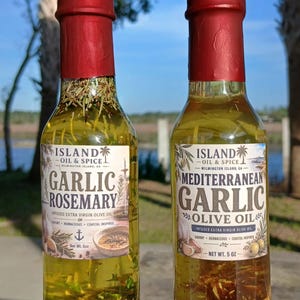 May include: Two glass bottles of infused olive oil with red wax seals. One is labeled "Garlic Rosemary" and the other "Mediterranean Garlic Olive Oil." Both are from Island Oil & Spice, Wilmington Island, GA. Each bottle contains 5 oz of oil.