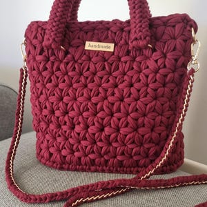 Puede incluir: Un bolso de mano de ganchillo burdeos hecho a mano con un patrón floral texturizado. El bolso tiene un asa trenzada a juego y una correa para el hombro desmontable con detalles de cadena dorada. Una pequeña etiqueta de madera dice "handmade".