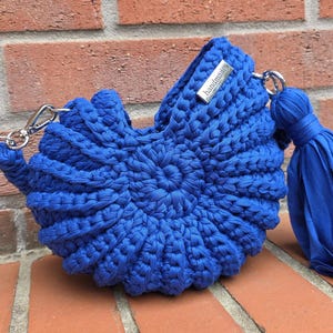 Könnte beinhalten: Eine leuchtend königsblaue Häkeltasche mit einem kreisförmigen, muschelartigen Strukturmuster. Die Tasche verfügt über ein silberfarbenes Metallschild mit der Aufschrift "handmade" und eine passende blaue Stoffquaste an einem silbernen Verschluss.