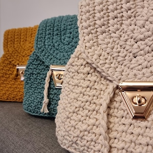 以下が含まれることがあります： マスタードイエロー、ティール、クリーム色の手作りのかぎ針編みバッグ3つ。各バッグは、ゴールドトーンの留め金が付いたフラップクロージャーが特徴です。バッグは厚く、質感のあるステッチで作られており、灰色の表面に表示されています。