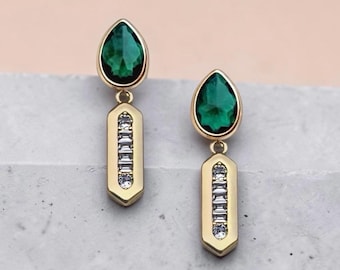 May include: A pair of gold-toned drop earrings. Each earring features a teardrop-shaped, emerald-green gemstone at the top, connected to a rectangular pendant with a row of small, clear crystals. The earrings are displayed on a light gray surface.