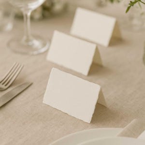 May include: Three off-white place cards with textured edges are arranged on a beige linen tablecloth. A fork, knife, and wine glass are visible in the background, suggesting a formal dining setting. The cards are folded to stand upright.