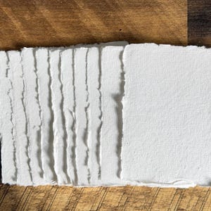 May include: A stack of handmade, white paper sheets with deckled edges. The paper has a textured surface and is arranged on a wooden background. The paper is likely used for art, calligraphy, or journaling.