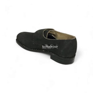 May include: Black suede shoe with a rounded toe and a strap across the top. The shoe has a low heel and a stitched sole. The text "Velikor Supply" is visible on the side of the shoe.