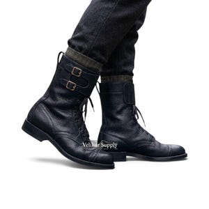 May include: A pair of black leather mid-calf boots featuring a textured surface, a cap toe design, and two adjustable straps with metal buckles across the upper shaft. The boots include laces and are worn with dark grey trousers and olive green socks.
