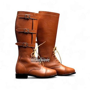 May include: A pair of brown leather riding boots. One boot features three buckle straps, while the other has laces. Both boots have a rounded toe and a small heel. The boots are set against a white background.