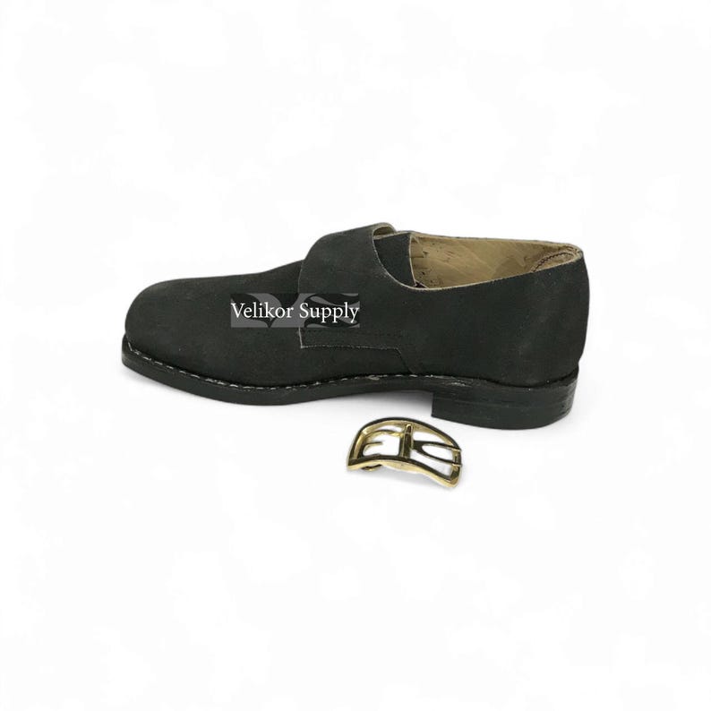 May include: A black leather shoe with a buckle, likely for historical reenactment or costume use. The shoe has a strap across the top and a stitched sole. A gold-colored buckle is separate.