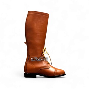 May include: A tall, brown leather riding boot with a cap toe and a small heel. The boot features a lace-up design with yellow laces and a buckle detail near the top. The text "Velikor Supply" is visible on the side of the boot.