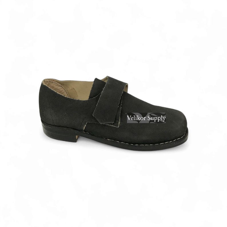 May include: Black leather shoe with a strap closure. The shoe has a rounded toe and a low heel. The text "Velikor Supply" is visible on the side of the shoe. The shoe is designed for comfort and durability.