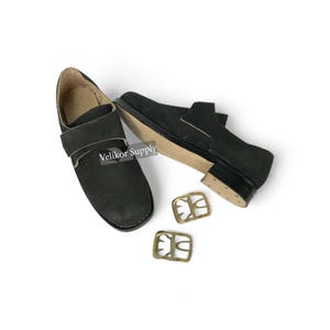 May include: Black leather shoes with a strap and buckle detail, alongside two gold-colored buckles. The shoes have a classic design, with a visible sole and heel. The text "Velikor Supply" is visible on the shoe.