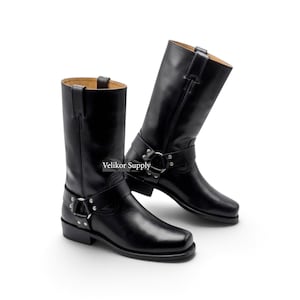 Könnte beinhalten: Ein Paar schwarze wadenhohe Stiefel mit einem Riemen, der mit einem silberfarbenen Metallring und Nieten versehen ist. Die Stiefel haben eine quadratische Zehenpartie, einen niedrigen Absatz und Zuglaschen. Der Text Velikor Supply ist sichtbar.